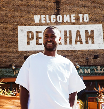 Clement, a Black man with short hair and a beard is standing in front of the words Welcome to Peckham painted on a wall